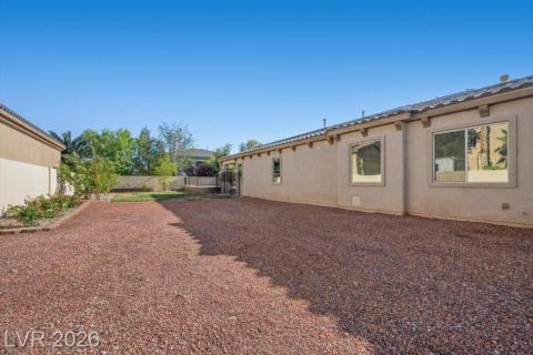 A spacious, low-maintenance backyard awaits, featuring a gravel surface and thriving shrubbery under a clear sky.